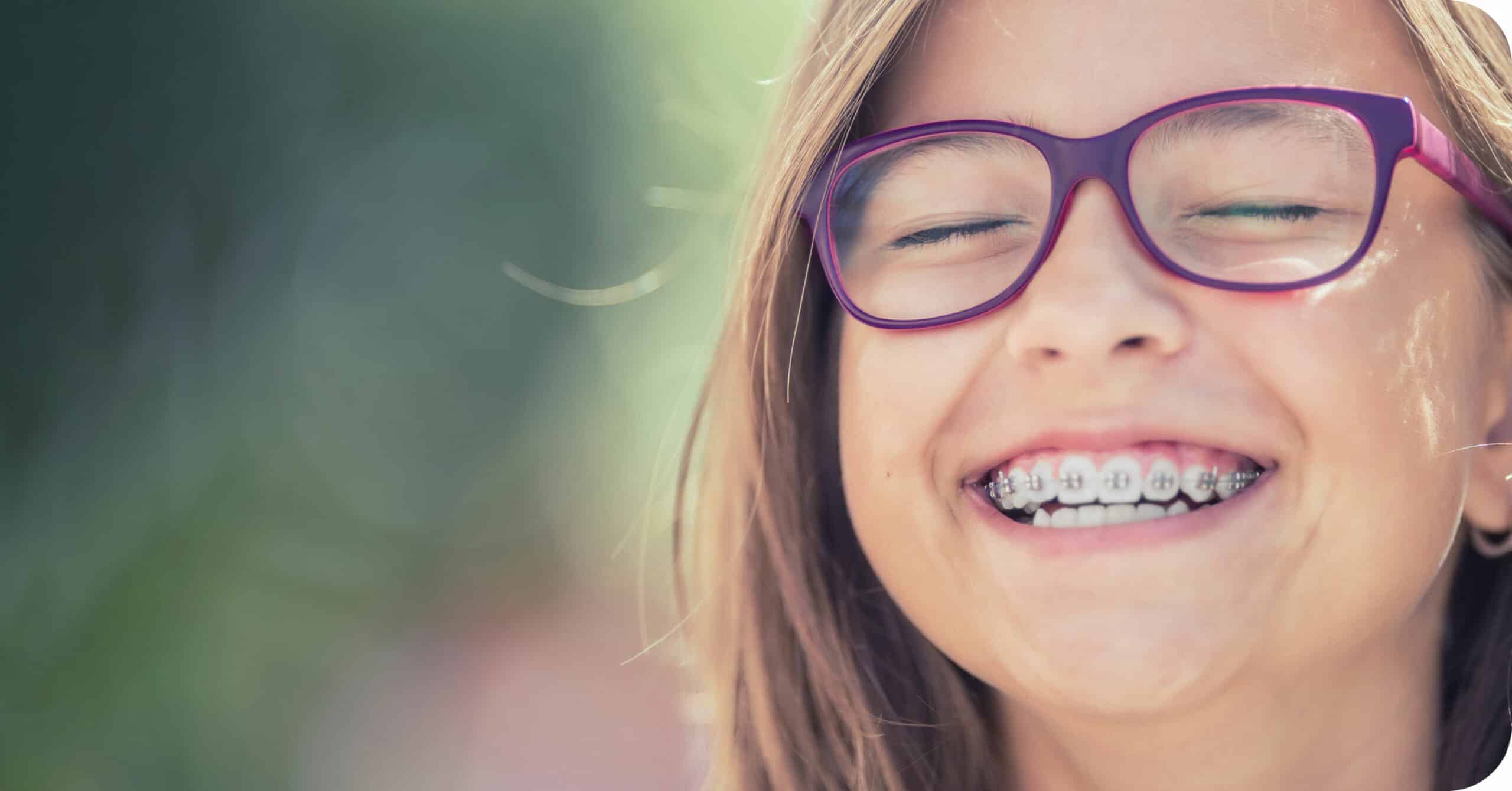 A child with glasses and braces is smiling brightly outdoors with a blurred greenery background. No recognizable landmarks or historical buildings are visible.